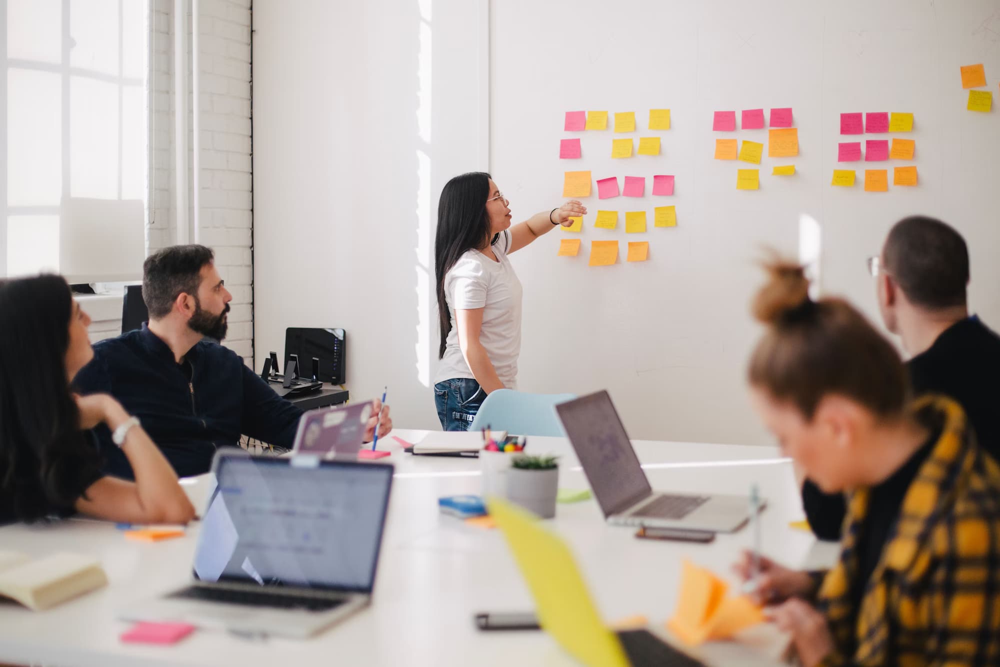 Atelier de travail : une personne désigne des notes autocollantes affichées au mur, face à des collègues installés autour d'une table avec leurs ordinateurs.